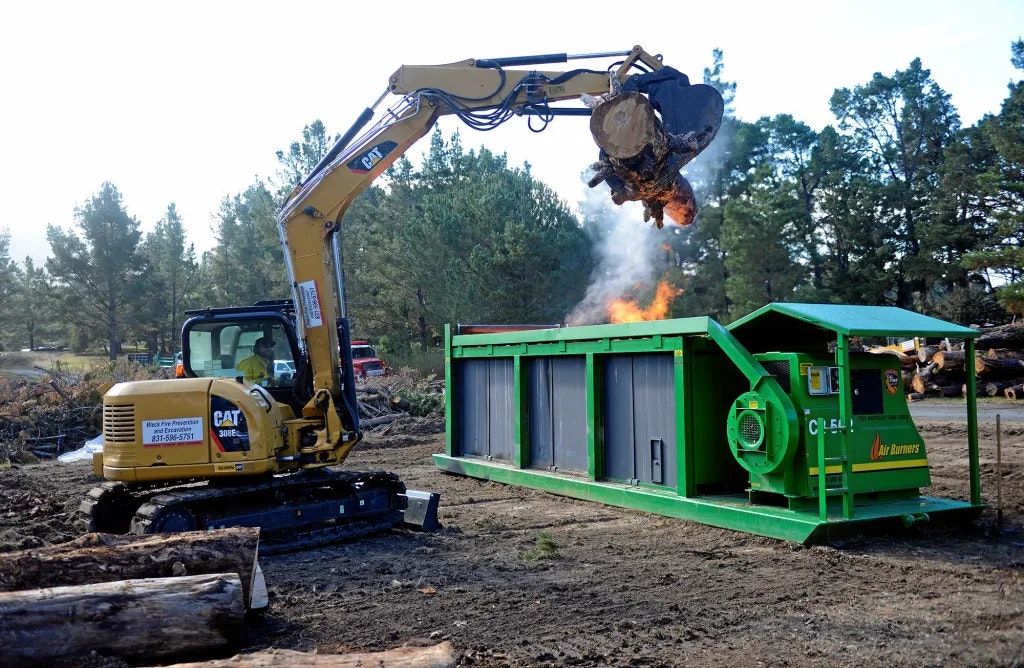 Excavator loads burning log into air curtain incinerator