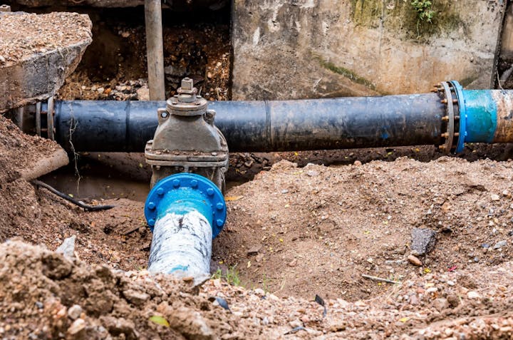 A dug-out area showing large black and blue pipes and a valve, likely part of a plumbing or water supply system.