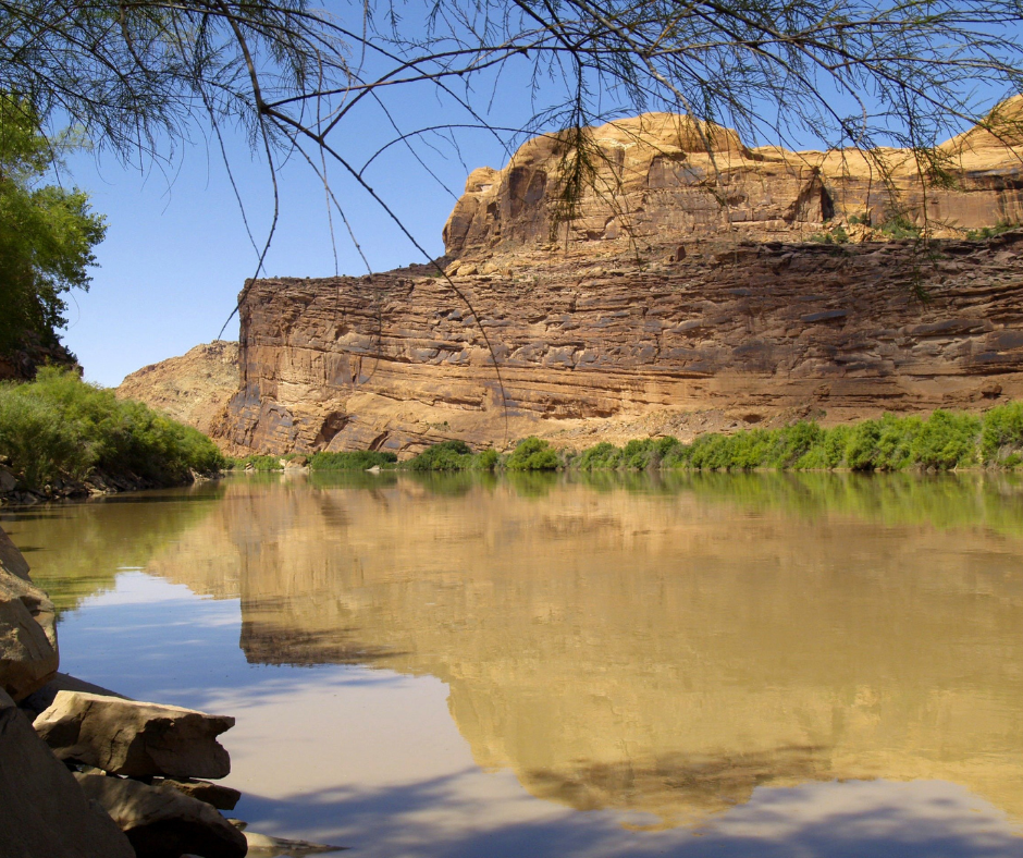 A tranquil river reflecting rugged cliffs under a clear blue sky, with green shrubs and rock formations.