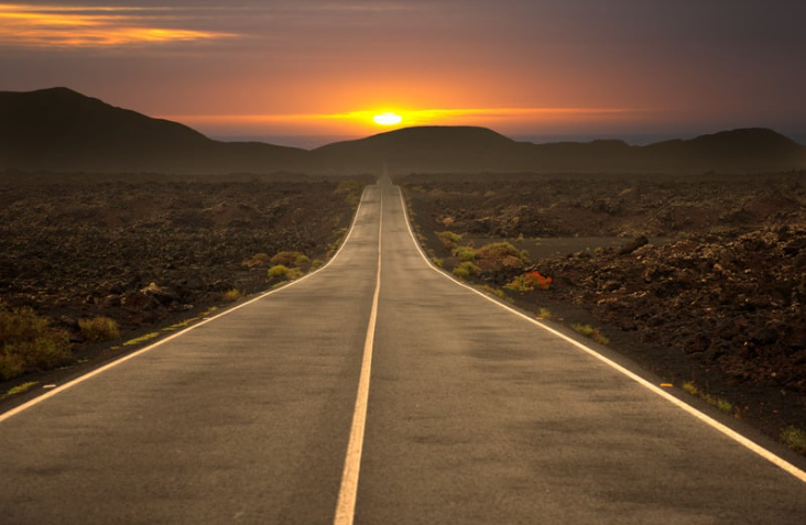 A straight empty road stretches into the horizon, with a stunning sunset illuminating the sky and surrounding volcanic landscape.