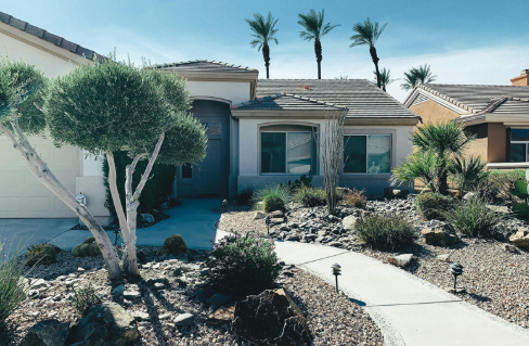 The image shows a modern house with a landscaped front yard featuring rocks, desert plants, and palm trees under a clear sky.