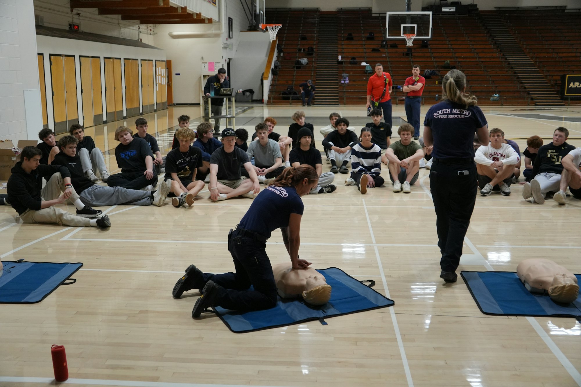 A CPR training session in a gym, with an instructor demonstrating on a dummy while students observe.