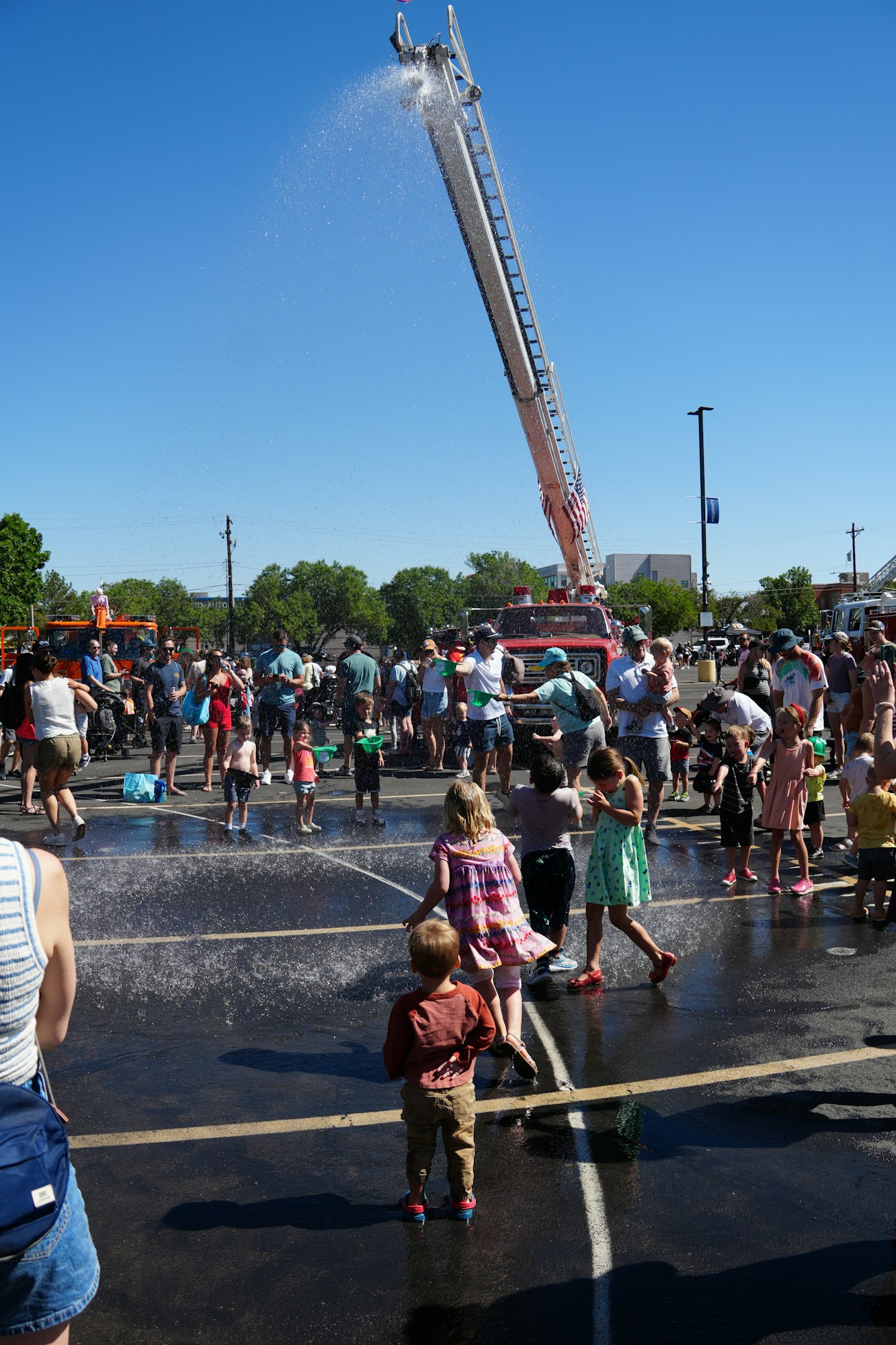 A lively scene with children and adults playing in water, enjoying a sunny day, with a fire truck spraying water in the background.