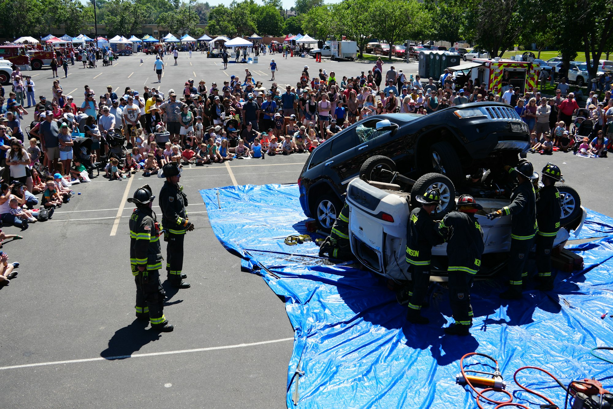 Firefighters demonstrate a vehicle recovery drill with an overturned car in front of a large audience at an outdoor event.