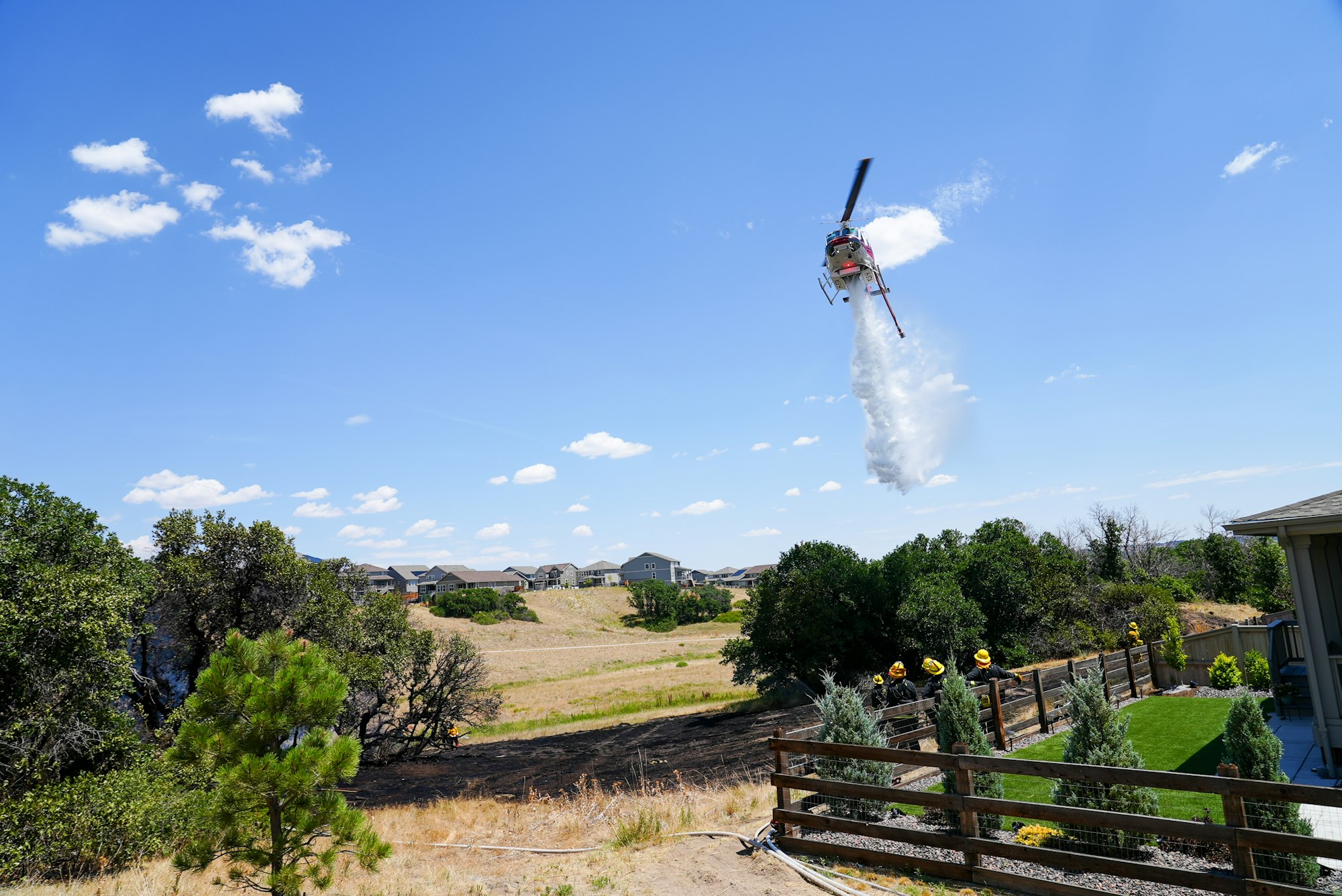 A helicopter is dropping water, likely for firefighting, over a dry landscape with houses and trees nearby.