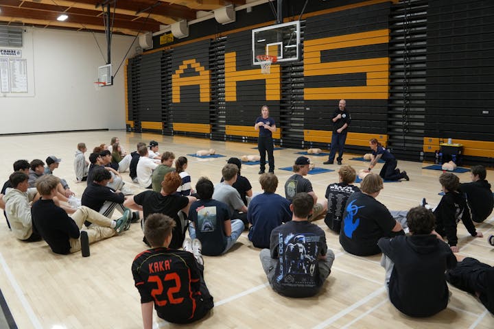 Students sitting in a gym watching a CPR training session with instructors and practice dummies.