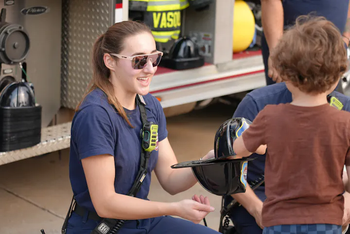 A firefighter interacts with a young child, showing them a helmet, with supportive figures in the background.