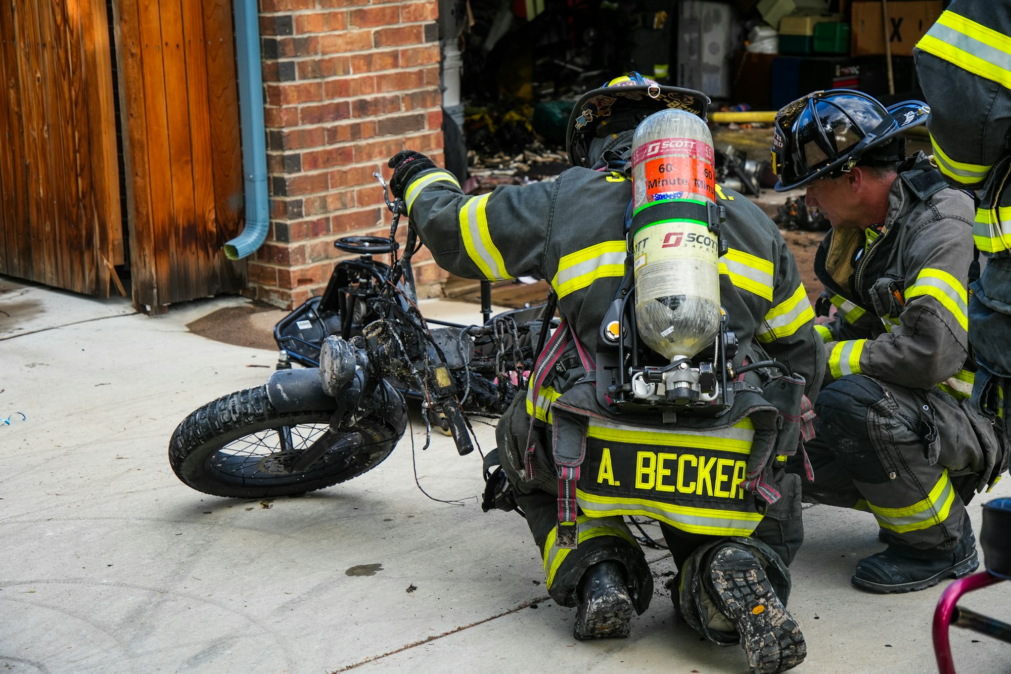 Firefighters assess a damaged motorcycle outside a garage, likely after a fire incident, wearing protective gear.