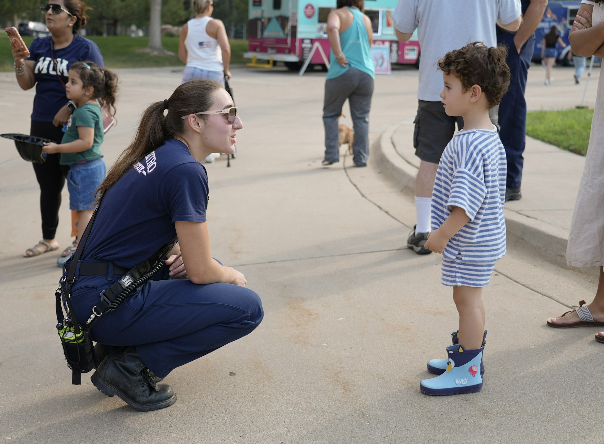 A child in striped clothes wearing colorful boots talks to a crouching woman in a uniform, with other people nearby.