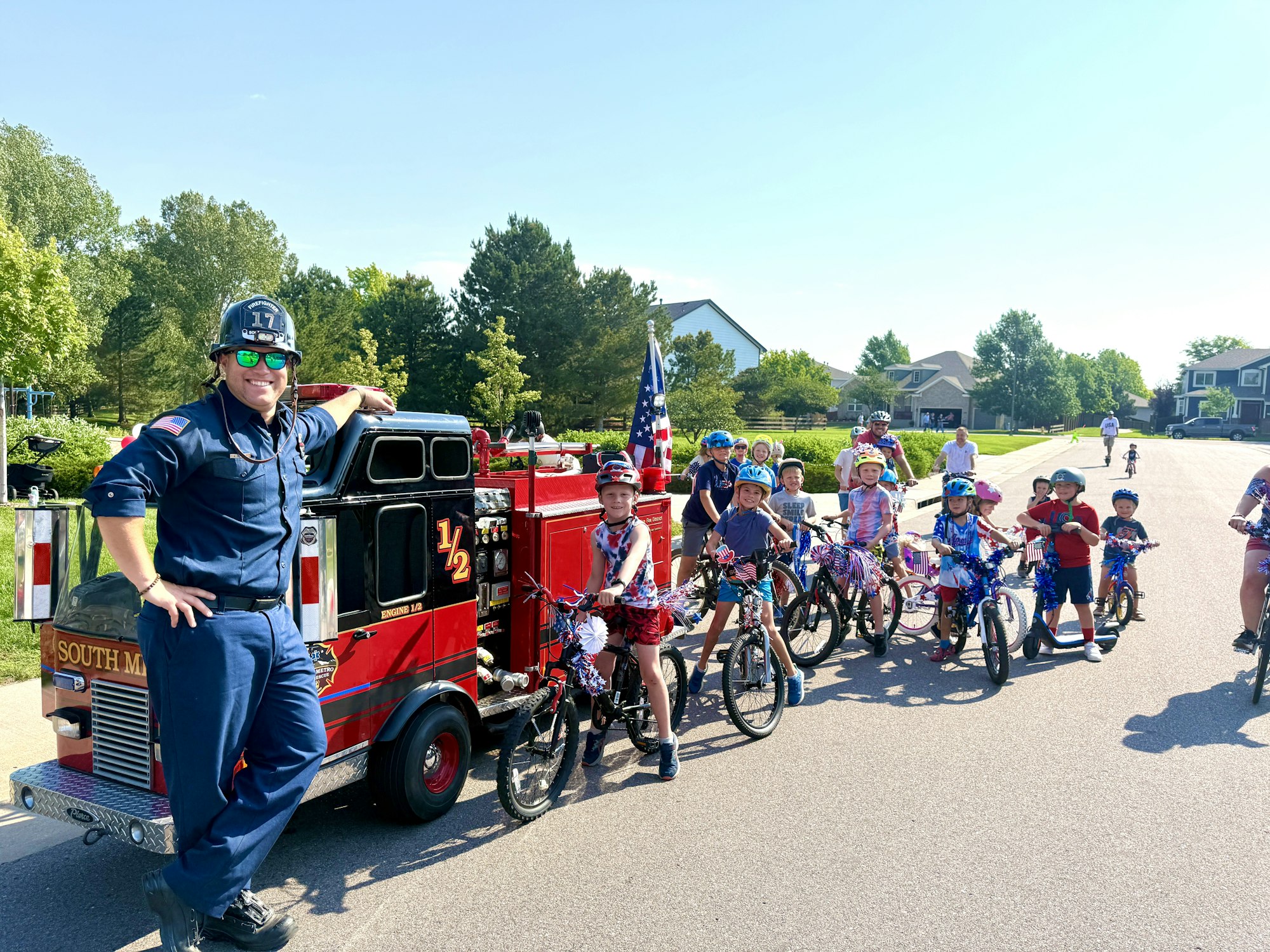 A firefighter poses with a group of children on decorated bikes, alongside a small fire truck in a festive neighborhood setting.