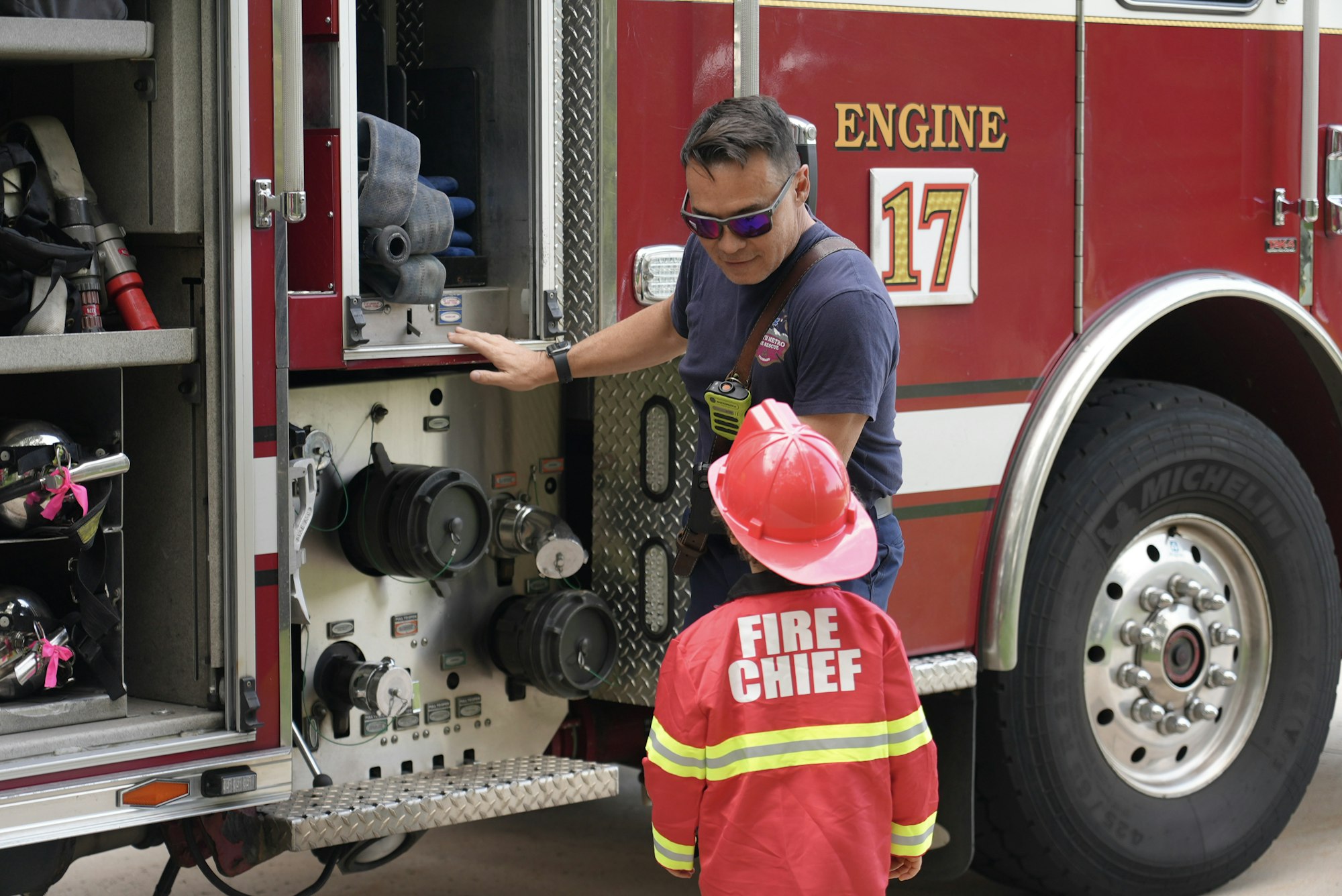 A firefighter shows equipment to a child in a fire chief costume at a fire truck.