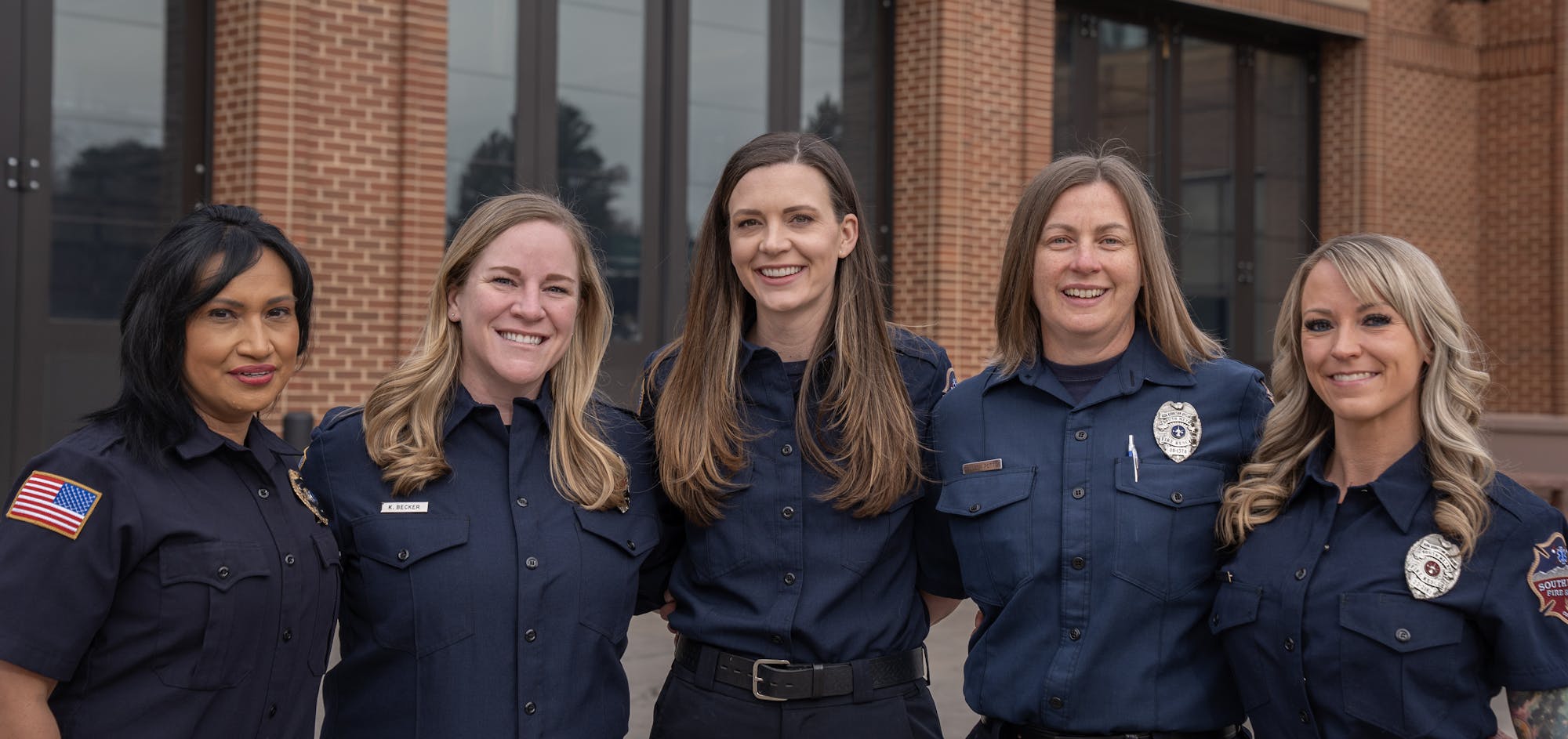 Five women in uniform, likely emergency responders, standing in front of a building with large windows.