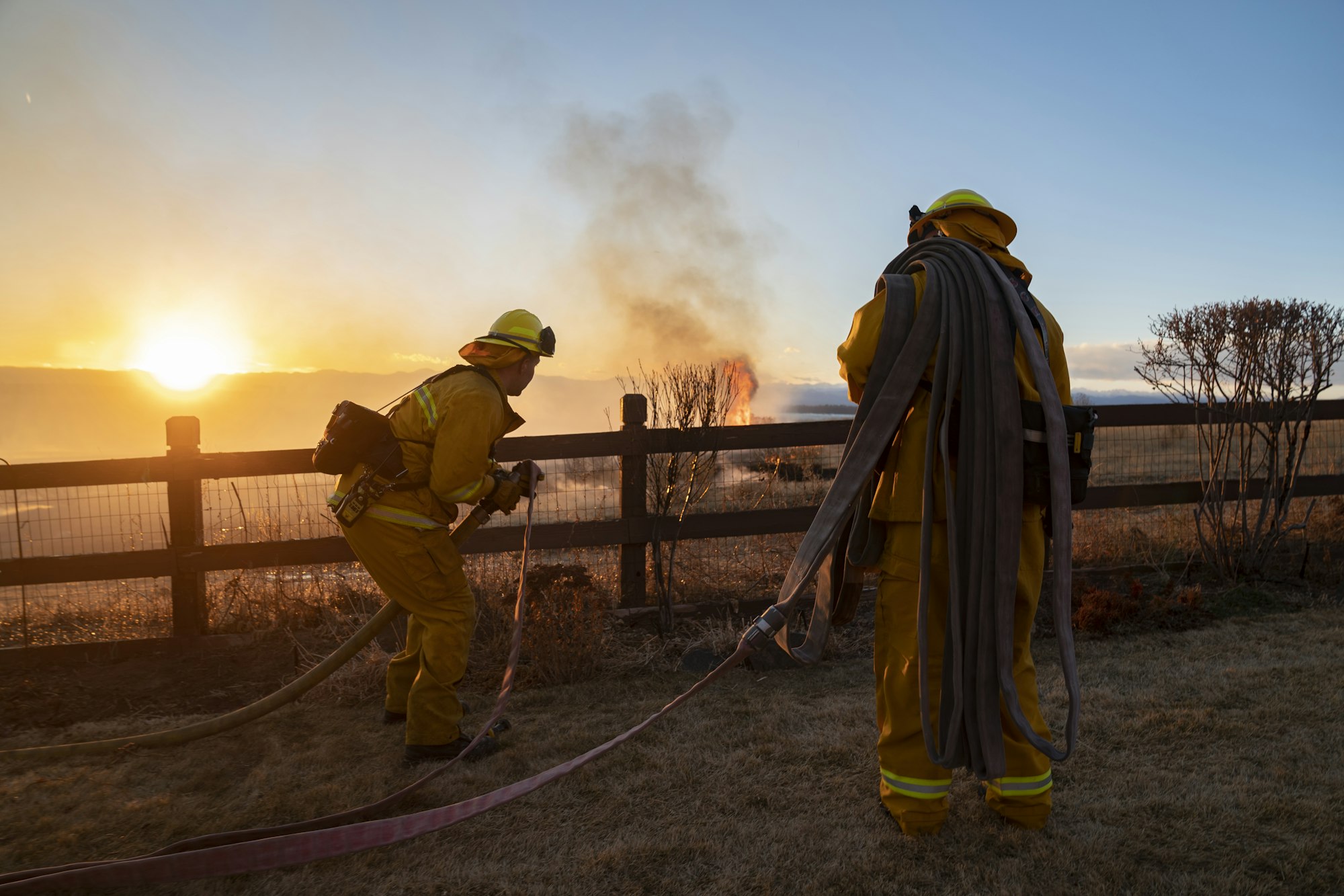 Two firefighters with hoses at sunset, preparing to tackle a fire.