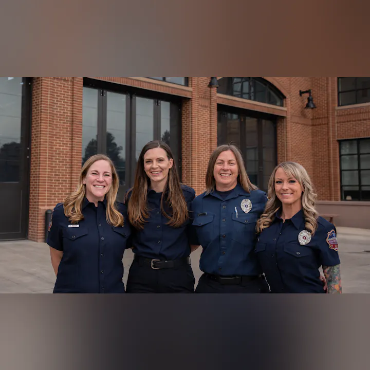 Four women in uniform, smiling together outside a building with large windows and brick walls.