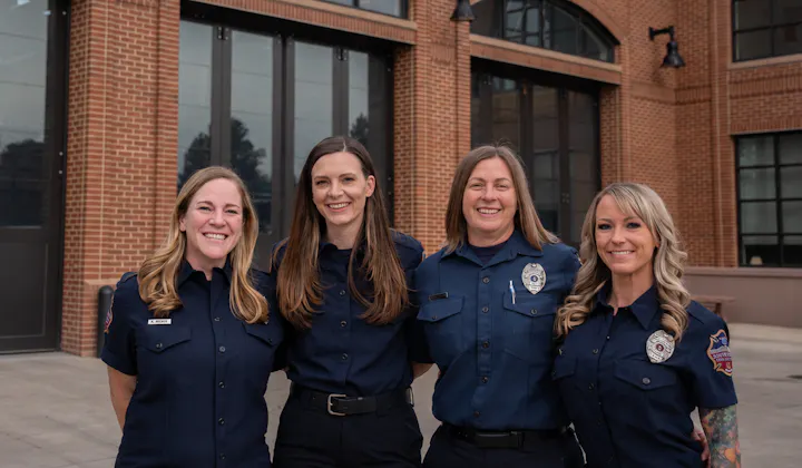 Four women in uniform, smiling together outside a building with large windows and brick walls.