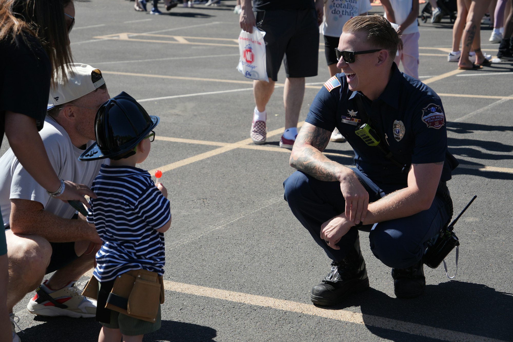 A police officer kneels and chats with a young boy wearing a toy firefighter hat and holding a lollipop in a sunny outdoor setting.