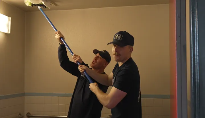 Two men are using a long pole to reach something on the ceiling, appearing to work together in a playful manner.