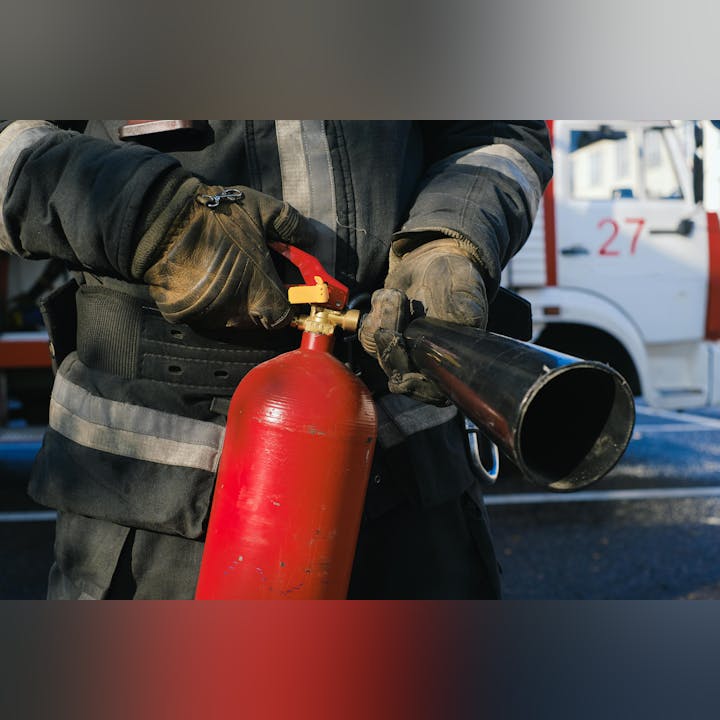 A firefighter in gloves holds a red fire extinguisher next to a fire truck.