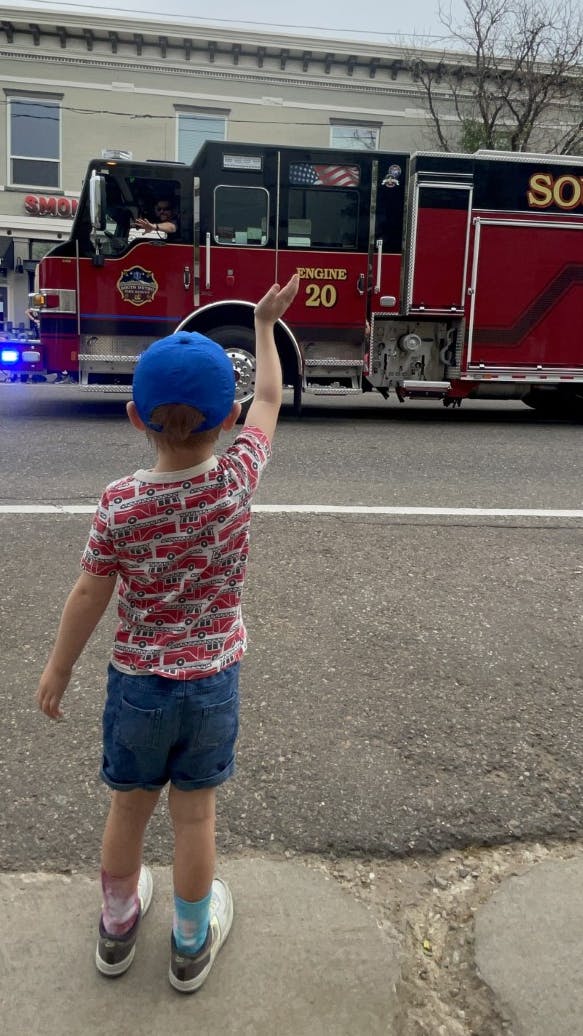 A child in a blue cap waves at a red fire truck labeled "Engine 20" on a street.
