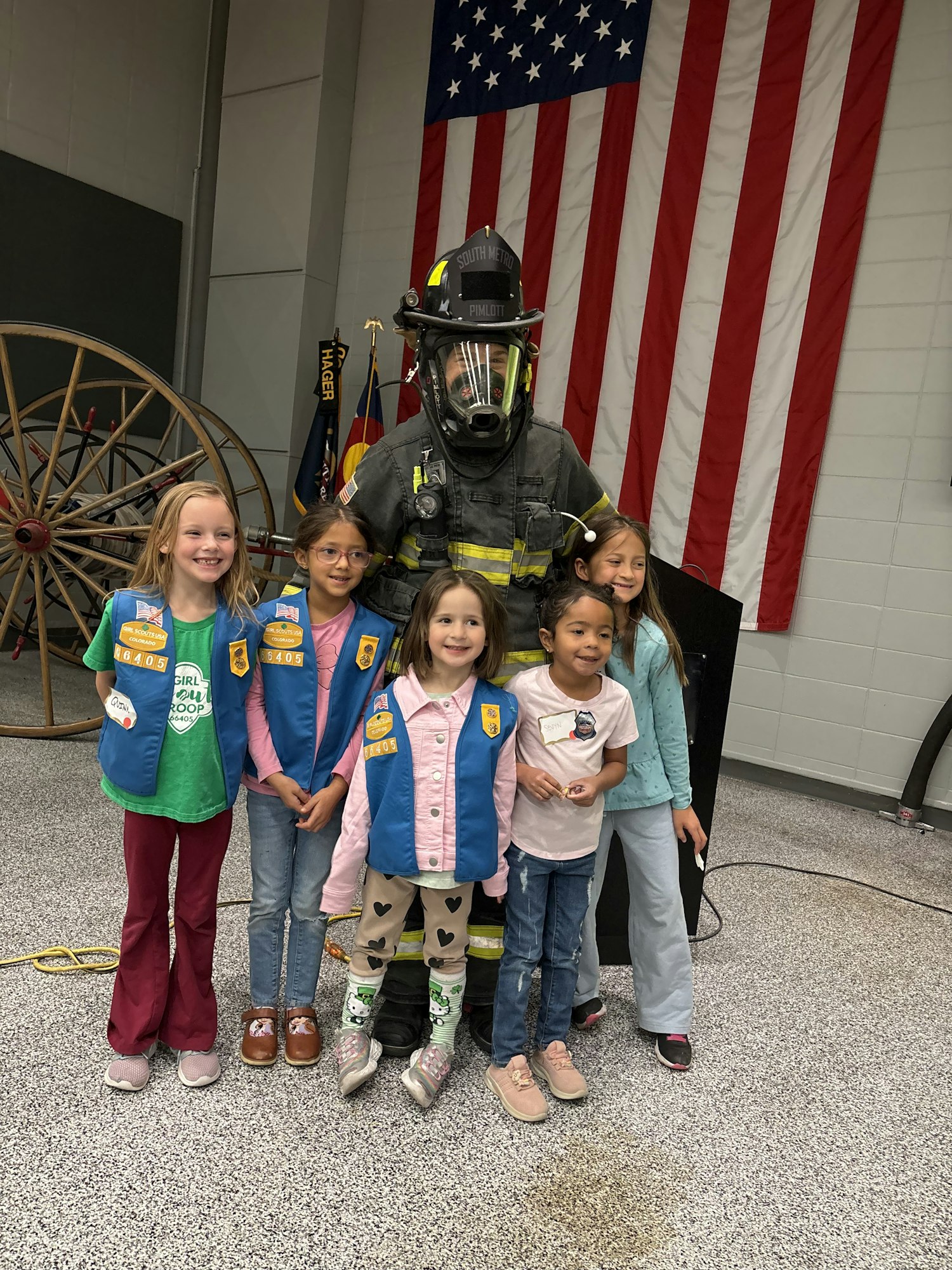 A group of five girls poses with a firefighter in gear, with an American flag backdrop and a vintage fire apparatus in the scene.