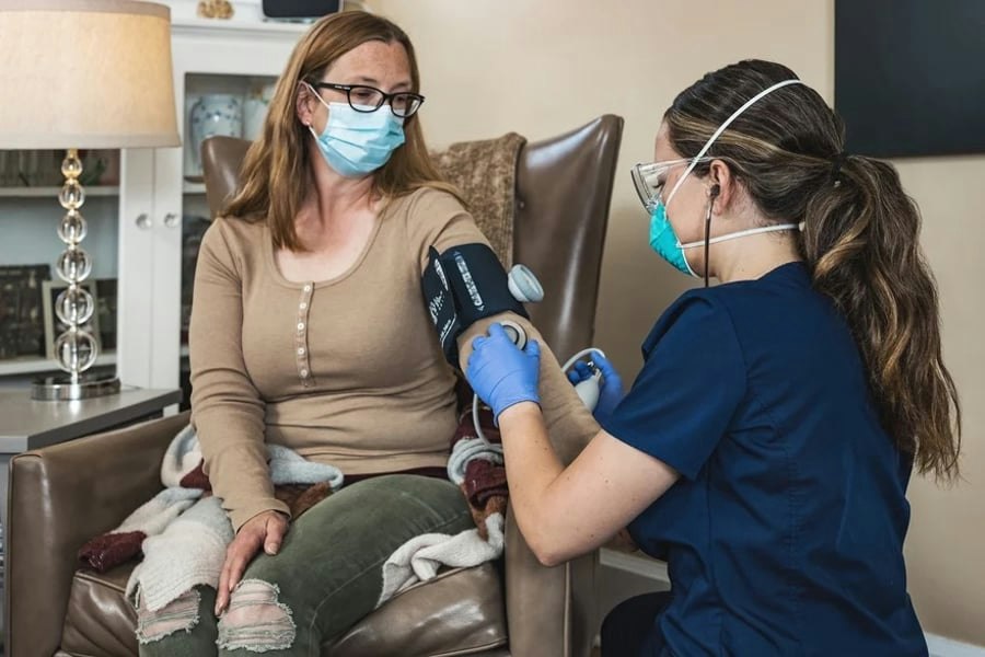 A healthcare worker in scrubs is checking the blood pressure of a masked woman sitting on a couch. They're both wearing masks.