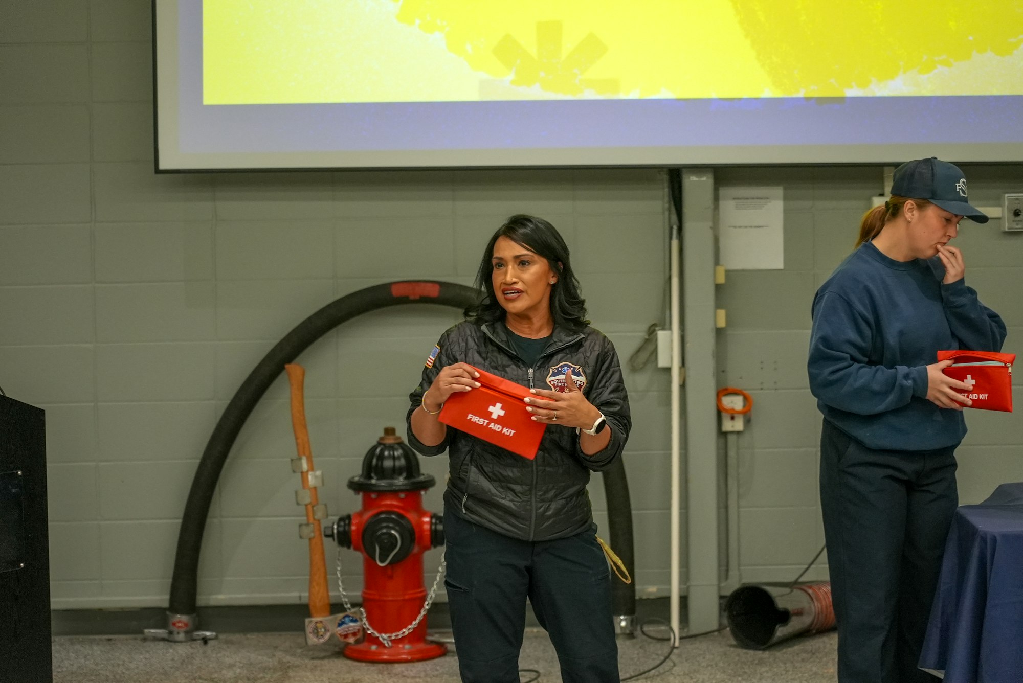 A woman is speaking about a first aid kit in a room with a fire hydrant and fire safety equipment nearby.