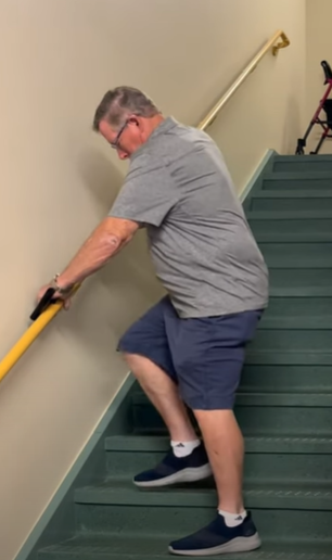 A person in casual clothing descends a staircase, using the handrail for support; a walker is visible at the top.