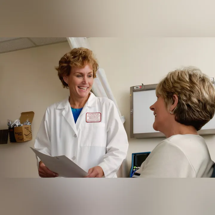 Two women smiling, one in a lab coat with a Urologic Oncology badge, holding papers in a clinical setting.