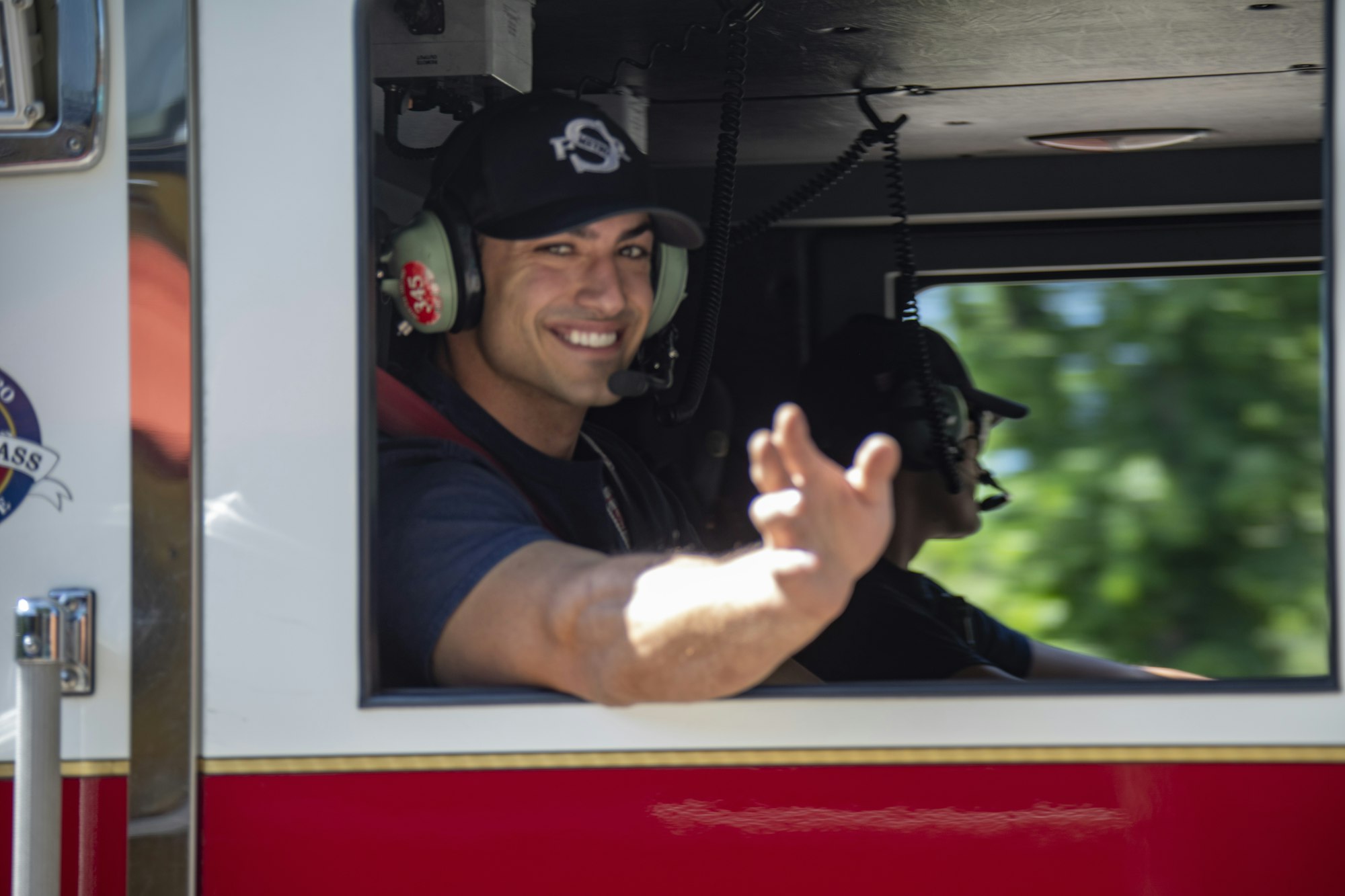 A smiling person in a fire truck is waving, wearing a headset, with another individual visible in the background.