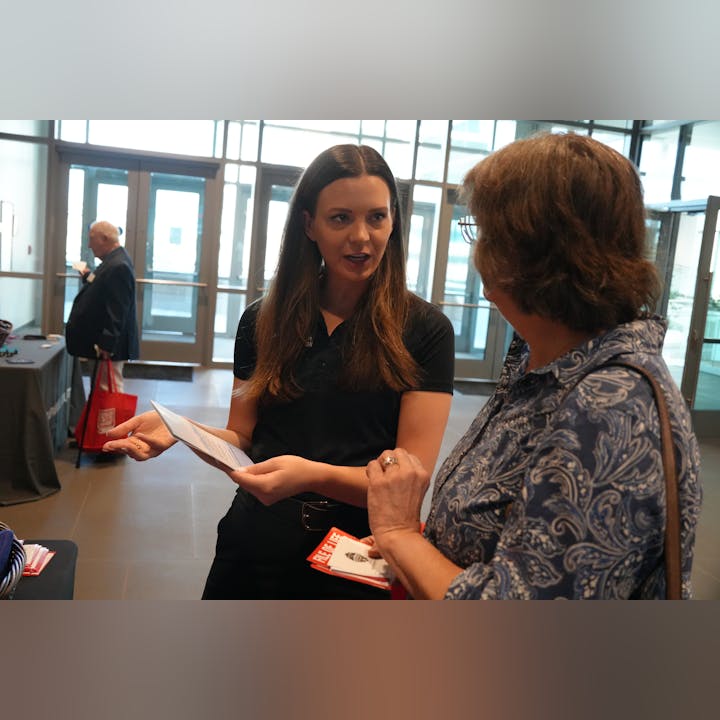 Two women engaged in conversation, one holding papers, indoors with people in the background.