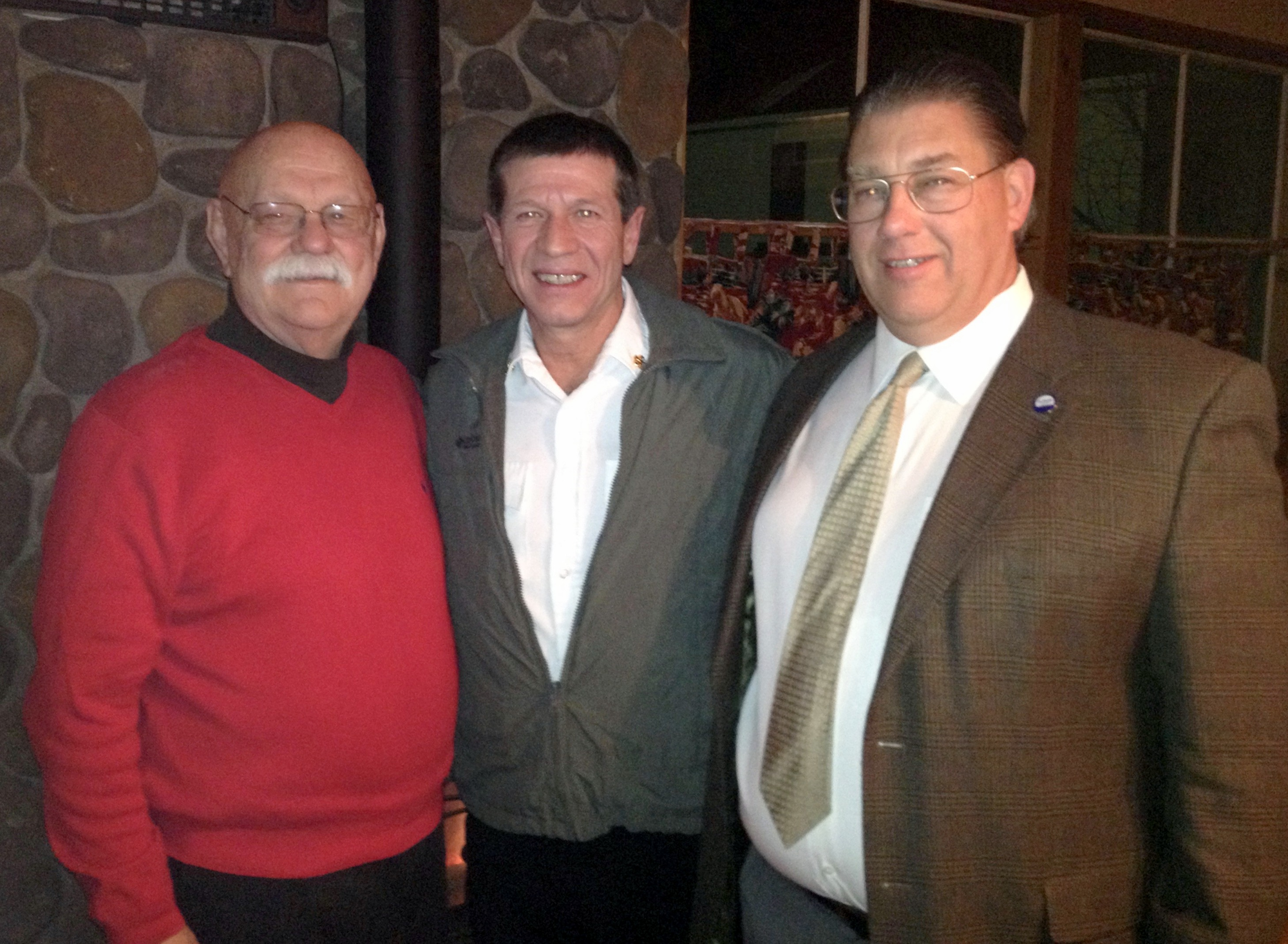 Three men standing together, smiling, in front of a stone wall indoors.