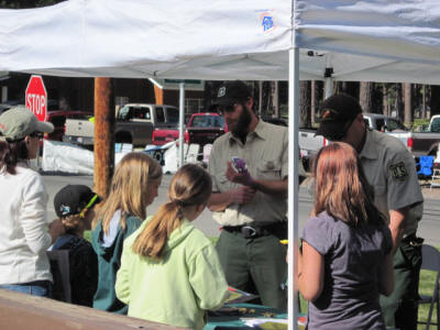 A group of kids engaging with a ranger under a canopy tent, with vehicles and trees in the background.