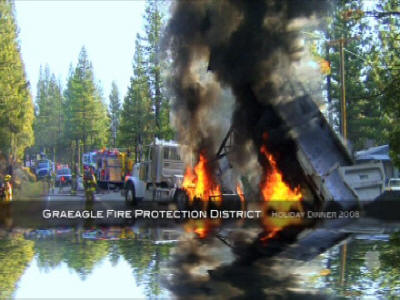 A burning structure with firefighters and a fire engine in the background, reflecting on water.