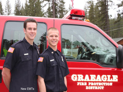 Two smiling EMTs in uniform standing in front of a red fire district vehicle.