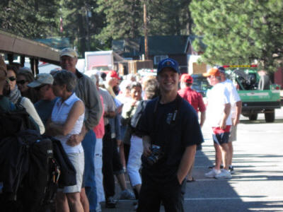 A person smiling in a cap, surrounded by a crowd of people, possibly at an outdoor event, with trees in the background.
