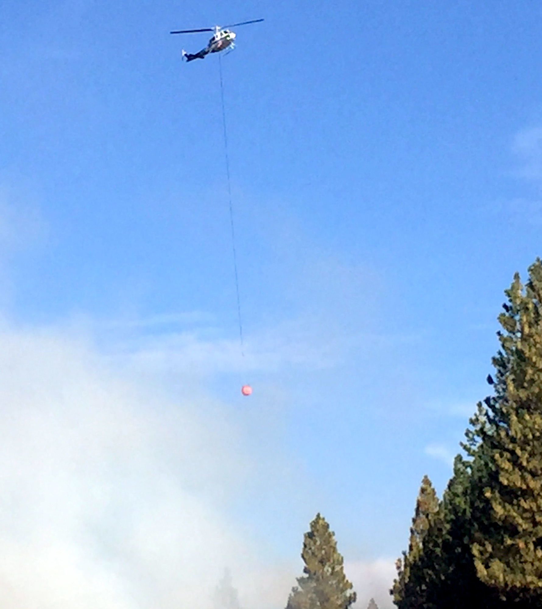 A helicopter with a long cable attached to a red object, flying over trees against a blue sky.