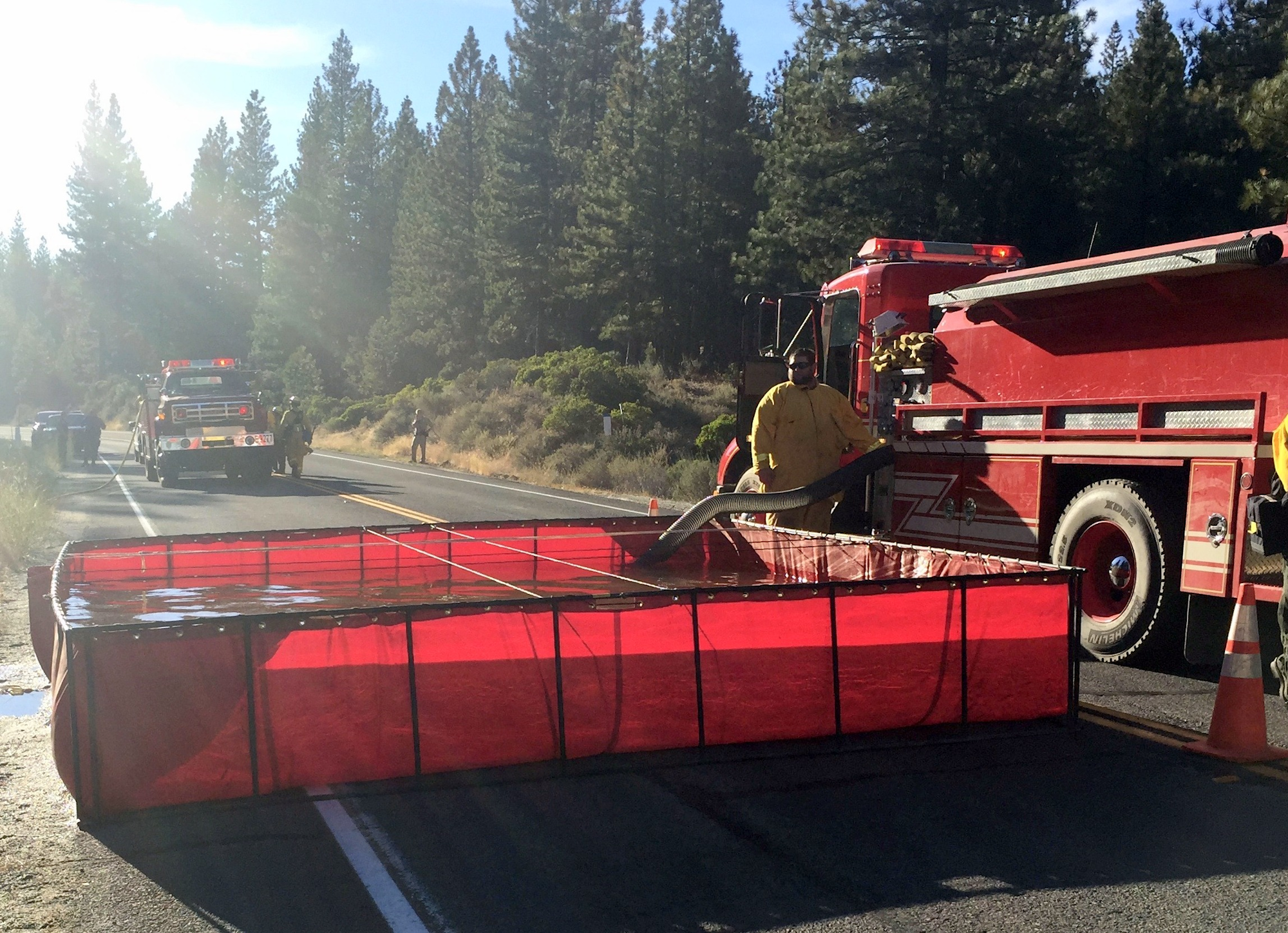 Firefighters filling a portable water tank near a fire truck on a sunlit road with trees in the background.