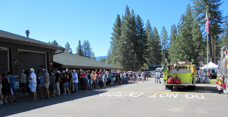 A long queue of people outside a building, a fire truck parked on the side, in a forested area with clear skies.