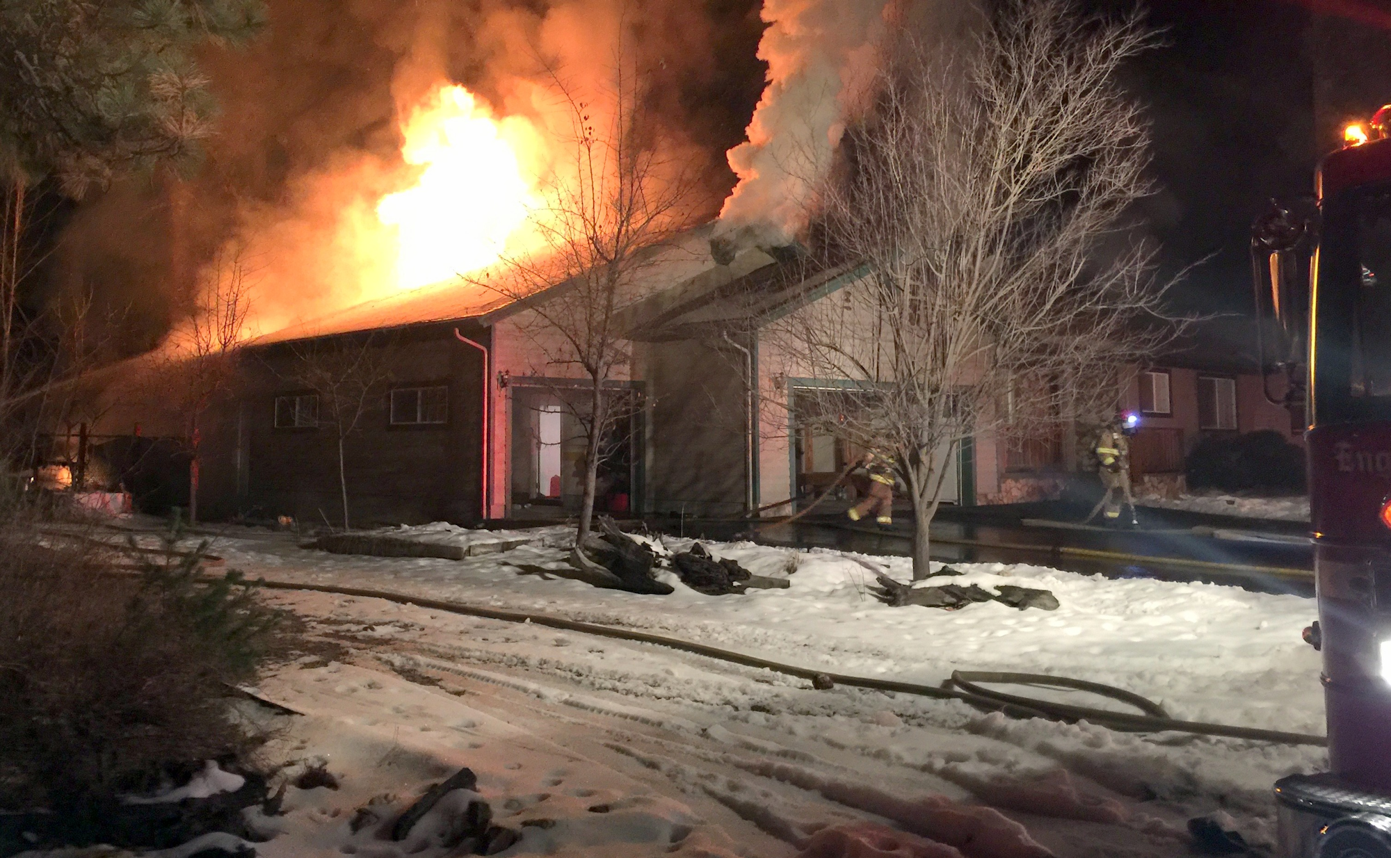 A house is engulfed in flames at night while firefighters work to extinguish the fire, with snow on the ground.