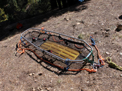 A stretcher, likely for mountain rescue or emergency services, with straps and carabiners on the ground.
