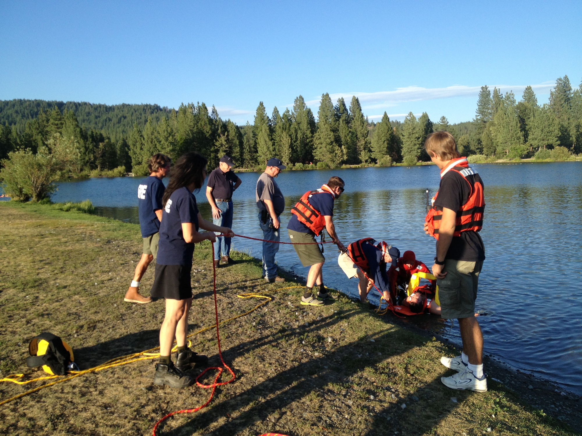 People in life jackets by a lake with rescue equipment, possibly a training exercise.