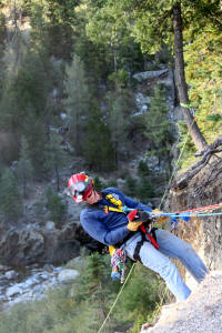 Person rappelling down a rock face with safety gear in a forested area.