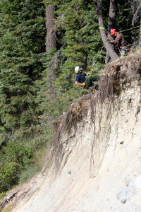 Two people wearing helmets on a steep, eroded slope with trees in the background. They appear to be involved in some kind of outdoor activity, possibly related to safety or training.