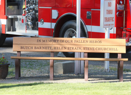 A memorial bench honoring fallen heroes Butch Barnett, Helen Strain, Steve Churchill with a fire truck in the background.