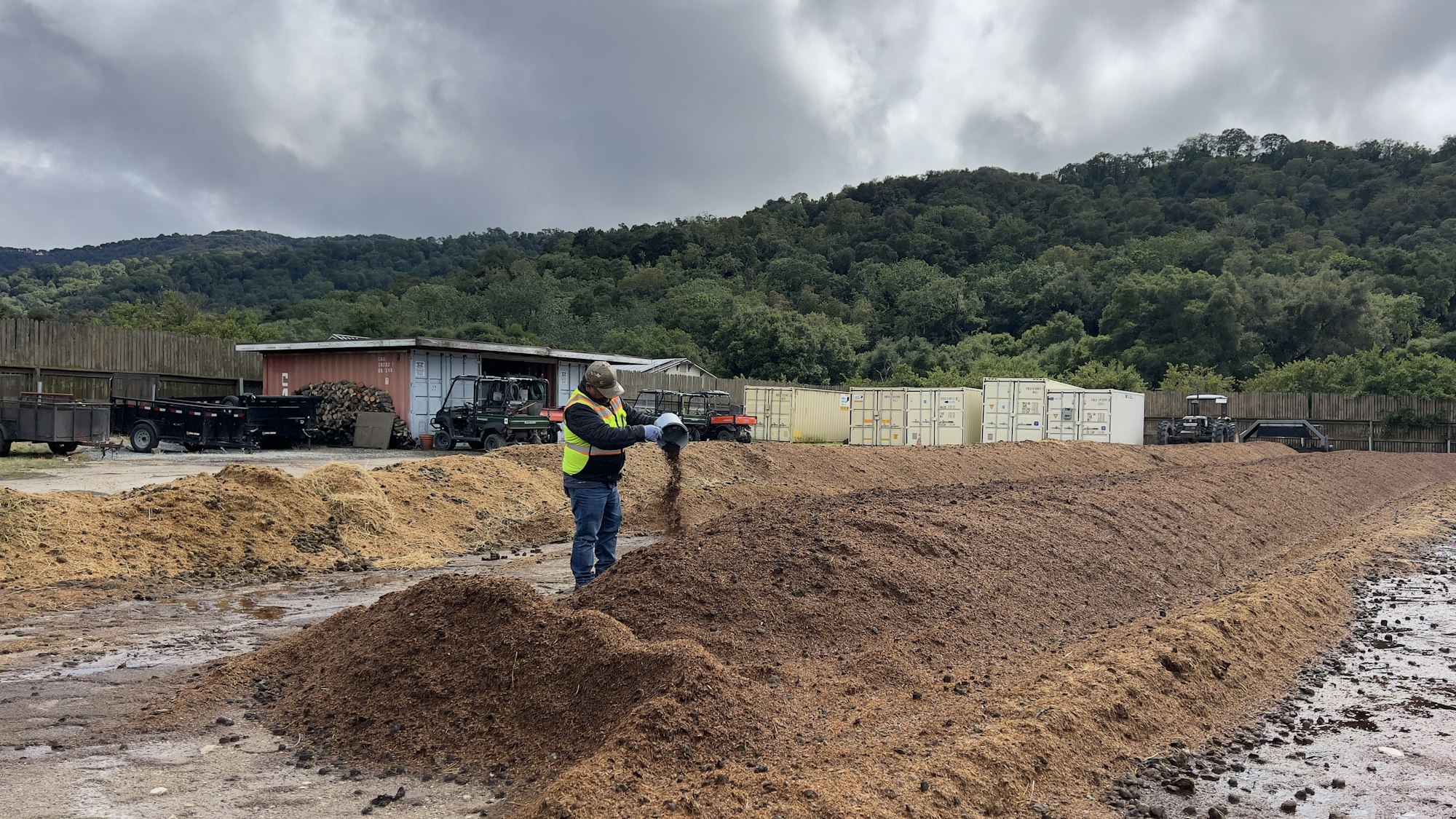 A worker in a safety vest spreads mulch or soil on a property with hills and storage containers in the background.
