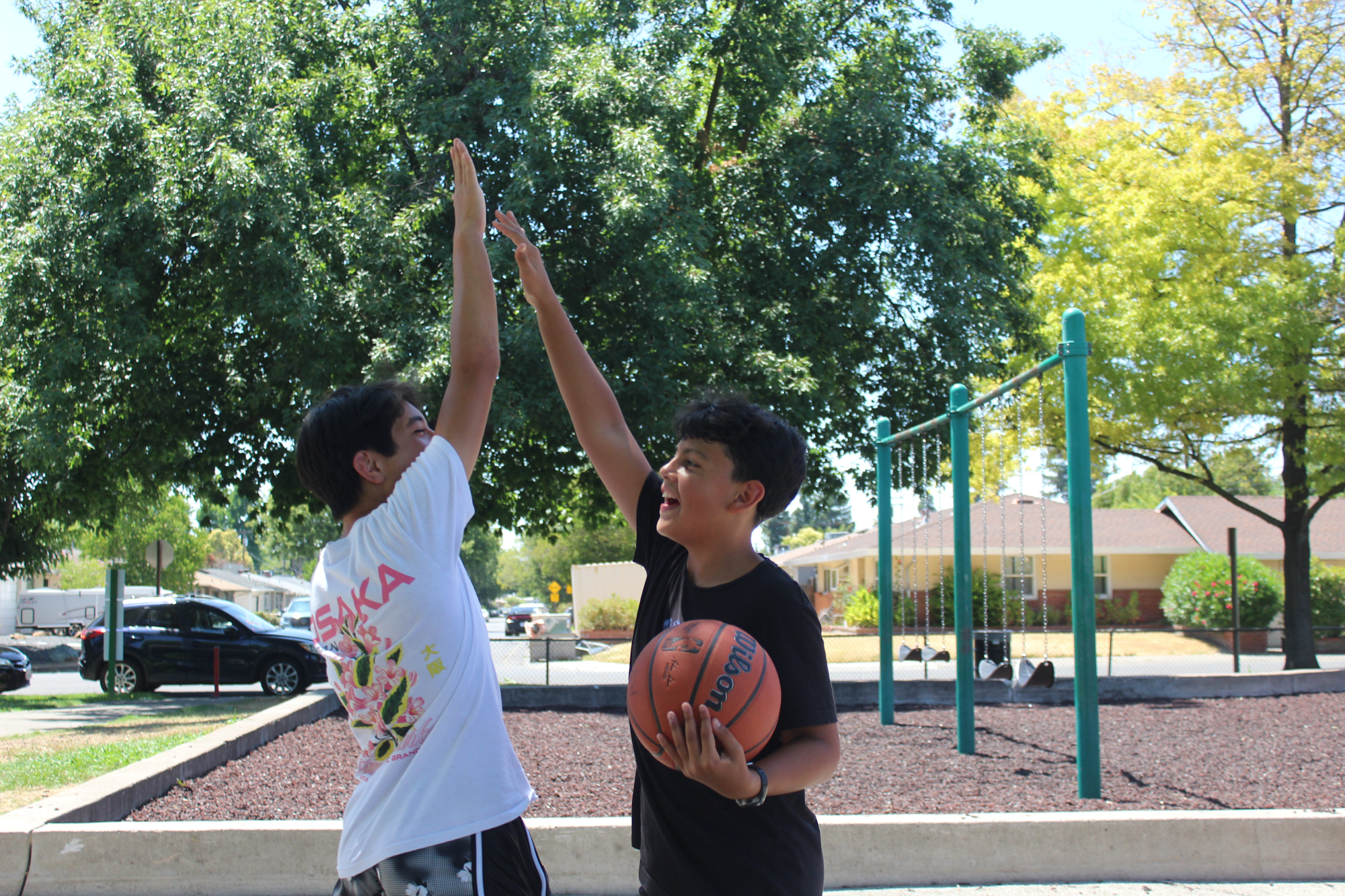 Two boys are high-fiving while playing basketball in a park on a sunny day, surrounded by green trees and playground equipment.