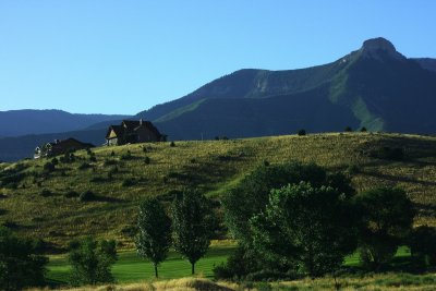 A scenic landscape featuring a house on a hill, surrounded by trees and grassy fields, with mountains in the background.