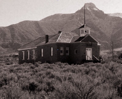 An old brick schoolhouse in a desolate area, with mountains in the background and surrounded by sparse vegetation.