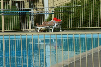 A person relaxes on a lounge chair by a swimming pool, wearing a red hat, surrounded by a gated area.