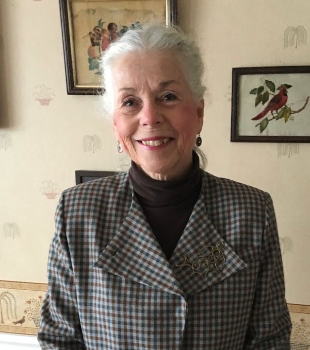 Ms. Beth Bigelow, BCLD Board Member with silver hair smiling, wearing a plaid blazer, standing in a room decorated with art on the wall.